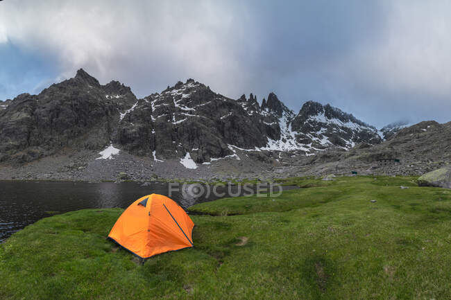 Tente orange située sur la rive herbeuse du lac Laguna Grande contre la chaîne de montagnes Sierra de Gredos et ciel nuageux à Avila, Espagne — Photo de stock