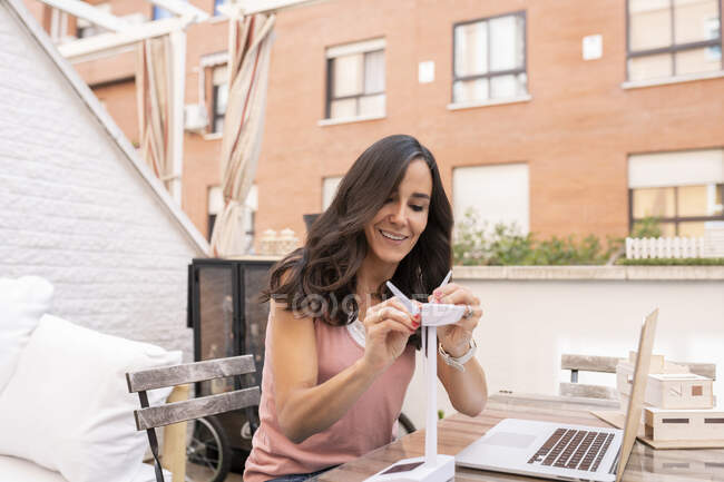 Arquiteta feminina feliz montando modelo de plástico de moinho de vento enquanto sentado à mesa com laptop e trabalhando no projeto no terraço — Fotografia de Stock