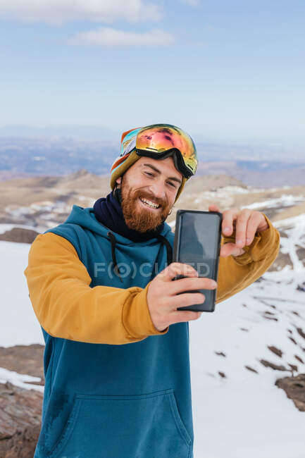 Esportista adulto com sorriso de dente e óculos esportivos tomando auto-retrato no celular contra cume nevado na Espanha — Fotografia de Stock