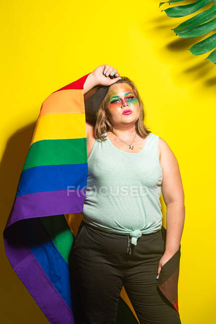 Overweight female with creative makeup holding rainbow flag and touching head against yellow background — Stock Photo