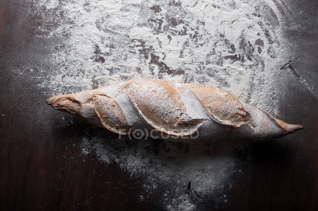 Appetitlich frisch gebackenes Baguette mit knuspriger Kruste auf Holztisch mit weißem Mehl vor schwarzem Hintergrund — Stockfoto
