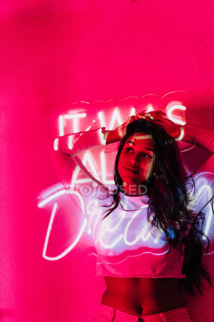 Junge hispanische Frau berührt Haare und schaut weg, während sie sich an Wand in der Nähe von Neon lehnt — Stockfoto