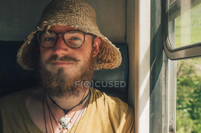 Sonriente hipster masculino en sombrero de paja viajando en tren en verano y mirando a la cámara cerca de las ventanas - foto de stock