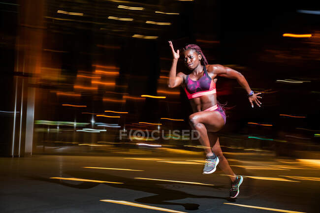 Vue latérale d'une femme sportive afro-américaine musclée courant dans une rue floue la nuit — Photo de stock