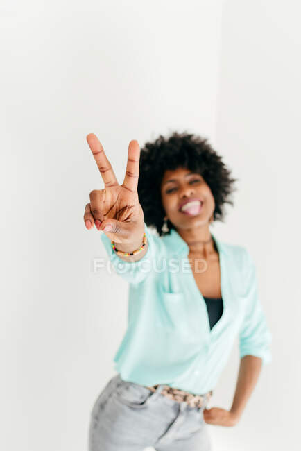 Playful young African American female in trendy outfit having fun and showing tongue and peace sign on white background — Stock Photo