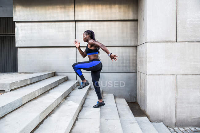 Vista lateral de una fuerte mujer afroamericana subiendo escaleras mientras entrena cerca de un edificio moderno en la calle de la ciudad - foto de stock