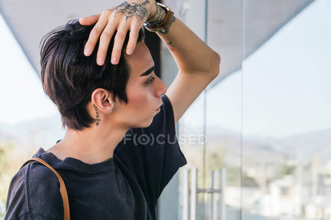 Seitenansicht eines stilvollen homosexuellen Mannes, der Haare berührt und in die Glaswand eines Gebäudes in der Straße blickt — Stockfoto