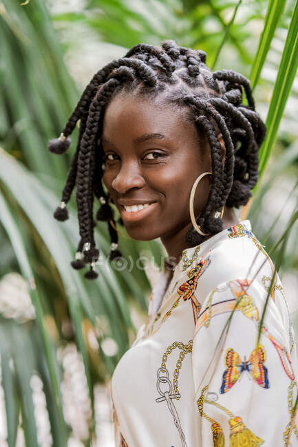 Stilvolle charmante schöne afroamerikanische Dame mit afrikanischen Zöpfen, die im grünen Park in die Kamera schaut — Stockfoto