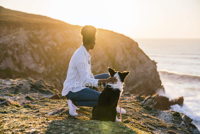 Seitenansicht der jungen afroamerikanischen Besitzerin mit Border Collie Hund, der die Zeit zusammen am Strand in der Nähe des winkenden Meeres bei Sonnenuntergang mit Blick auf die Landschaft verbringt — Stockfoto
