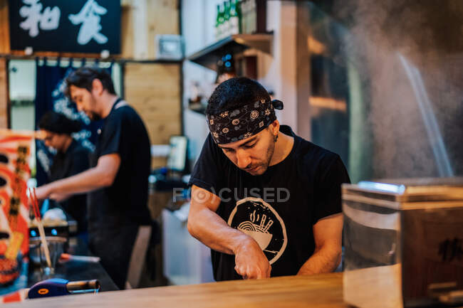 Young man in apron cooking Asian dishes while standing at counter in ramen bar — Stock Photo