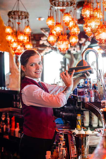 Side view of female barkeeper in uniform shaking cocktail in metal shaker while standing at counter in pub and looking at camera — Stock Photo