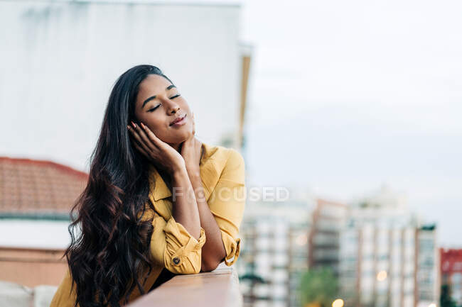 Молода іспаномовна жінка в повсякденному одязі спирається на поручні з закритими очима, відпочиваючи на балконі ввечері в місті. — стокове фото