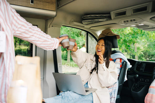 Cheerful young Asian woman with laptop clinking cup of coffee with unrecognizable friend inside camper van during summer journey in nature — Stock Photo