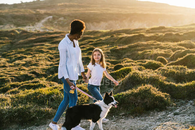 Full body side view of happy multiethnic woman and little girl with Border Collie dog walking together on trail among grassy hills in sunny spring evening — Stock Photo