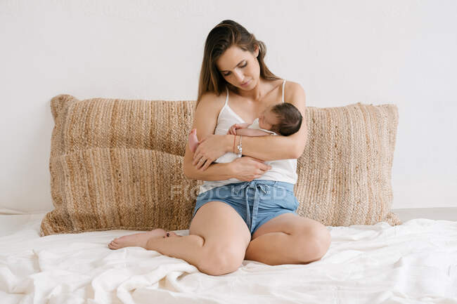 Zarte Mutter sitzt mit entzückendem Säugling auf weichem Bett im gemütlichen Zimmer zu Hause — Stockfoto