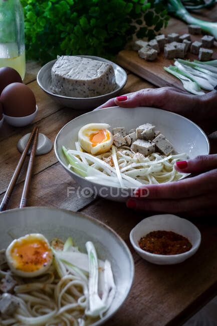 Сверху обрезанный неузнаваемый человек ест свежую вареную лапшу с тофу, яйца и овощи с палочками на деревянном столе — стоковое фото