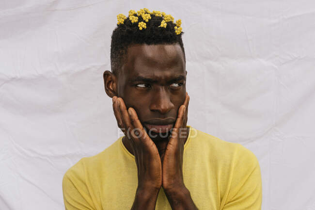 Bello afroamericano maschio con fiori gialli in capelli che toccano il viso e distogliendo lo sguardo su sfondo bianco — Foto stock