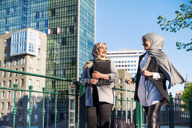 Mulheres empresárias muçulmanas sorridentes com mala andando pela rua na cidade e olhando umas para as outras — Fotografia de Stock