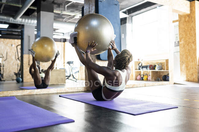 Atleta femenina afroamericana irreconocible en ropa deportiva que realiza ejercicio abdominal con pelota en forma en la estera contra el espejo en el gimnasio - foto de stock