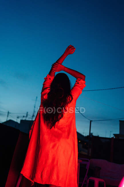 Vue arrière d'une femme ethnique méconnaissable en lingerie regardant la caméra tout en se reposant sur une terrasse sous la lumière rouge néon la nuit avec un ciel bleu foncé — Photo de stock