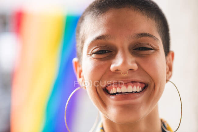 Joven mujer étnica soñadora con bandera colorida y pelo corto mirando a la cámara contra la pared sobre fondo borroso - foto de stock