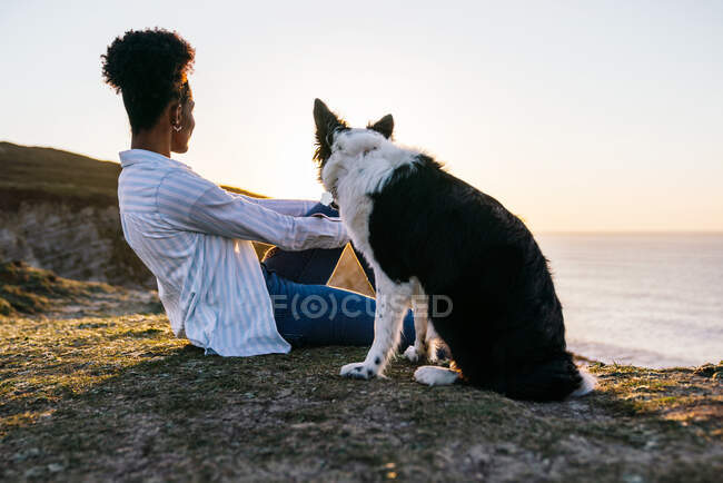 Vue latérale de la jeune propriétaire afro-américaine avec Border Collie chien passer du temps ensemble sur la plage près de la mer agitant au coucher du soleil contemplant des vues — Photo de stock