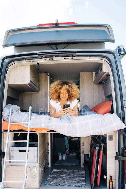 Fröhliche Afroamerikanerin mit lockigem Haar, lächelnd und Handy schmökernd, während sie sich während der Roadtrip im Wohnwagen auf dem Bett ausruht — Stockfoto