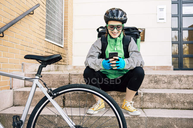 Full length happy delivery woman in uniform sitting on steps near bike and browsing cellphone while taking break during work — Stock Photo