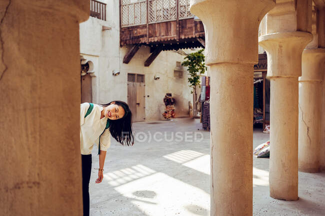 Happy young female tourist peeking from behind stone column of old building while visiting Al Fahidi Historical Neighbourhood in Dubai during summer holidays — Stock Photo