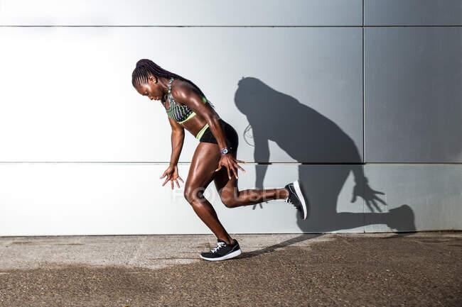 Мускулистая афроамериканка в спортивной одежде смотрит в камеру и прыгает во время тренировки на городской улице возле стены современного здания — стоковое фото