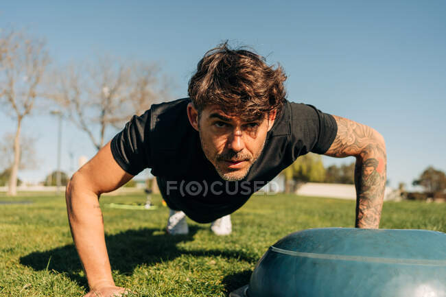 Nivel del suelo del atleta masculino determinado en entrenamiento de ropa deportiva en el prado mientras mira hacia adelante bajo el cielo azul - foto de stock
