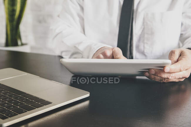 Crop empresario masculino anónimo en camisa blanca navegar por Internet en la tableta en el escritorio con el ordenador portátil durante el teletrabajo - foto de stock