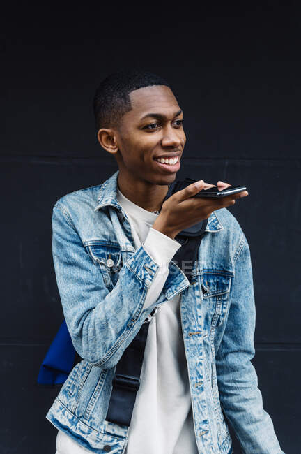Вид сбоку на молодого чернокожего человека с мобильным телефоном и рюкзаком на улице, оставляющего электронную почту — стоковое фото