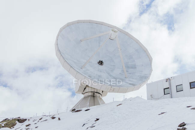 Desde abajo de la antena satélite en la montaña nevada contra la fachada del edificio bajo el cielo nublado en España - foto de stock