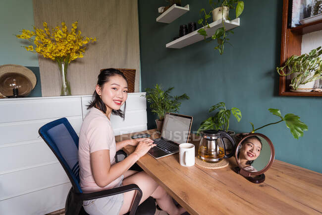 Vista lateral de feliz asiático freelancer feminino ler documentos no laptop enquanto sentado à mesa durante o trabalho remoto olhando para a câmera — Fotografia de Stock