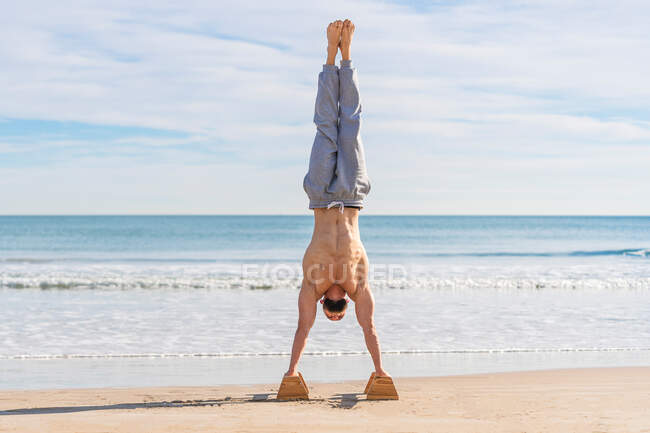 Full length of male athlete performing handstand with parallel bars on sandy coast with ocean waves on background — Stock Photo