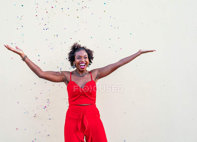 Возбужденная афроамериканка с распростертыми руками и открытым ртом, стоящая под падающими конфетти на белом фоне — стоковое фото