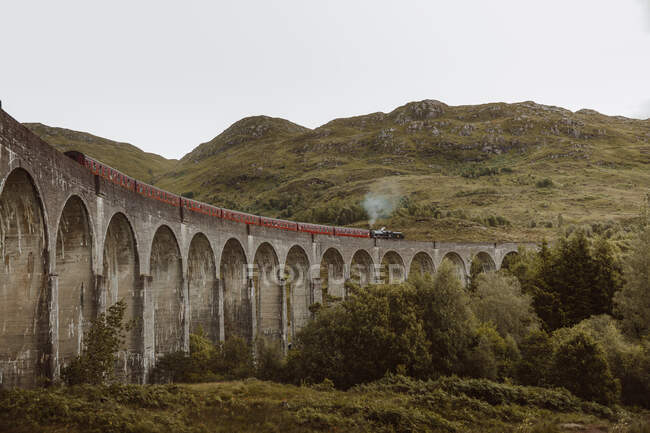 Steam train riding along old arch bridge near rough hill on gray day in Glenfinnan, UK countryside — Stock Photo
