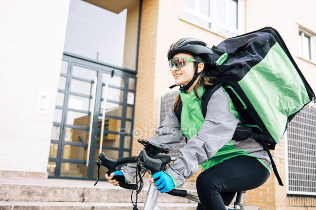 Низький кут жіночого кур'єра з термальною сумкою, посміхаючись і їздячи на велосипеді на вулиці, роблячи доставку в сонячний день у місті — стокове фото