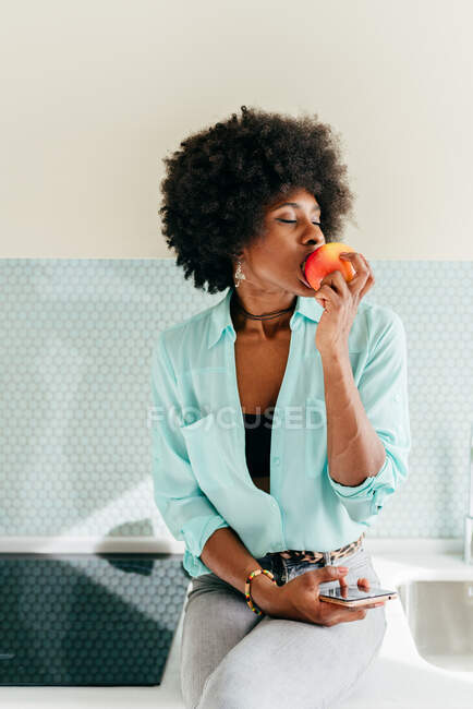 Belle femme afro-américaine moderne avec smartphone à la main assis sur le comptoir de cuisine avec les yeux fermés à la maison et manger de la pomme — Photo de stock