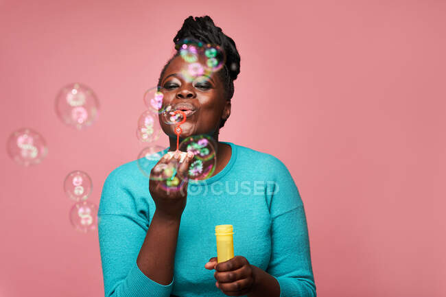 Felice femmina afroamericana guardando giù indossando vestiti blu e soffiando bolle di sapone contro sfondo rosa in studio — Foto stock