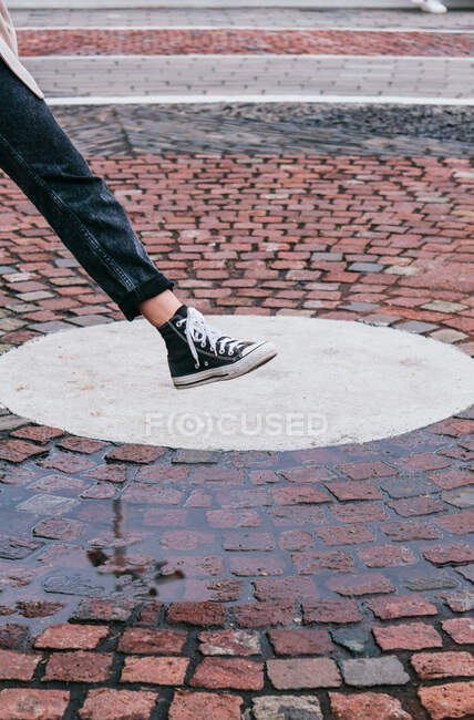 Mujer irreconocible en jeans elegantes y zapatillas de deporte pisando el charco en el pavimento mientras juega en la calle en el día lluvioso - foto de stock