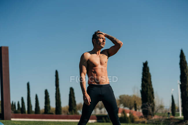 Мужчина без рубашки с татуировкой и широкими ногами тренируется, глядя на луг — стоковое фото