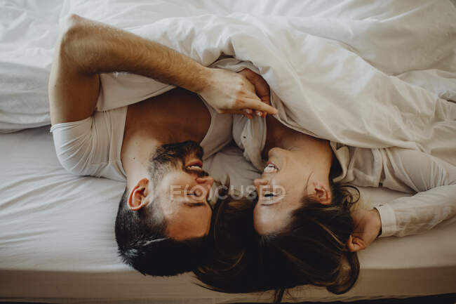 Jovem alegre e mulher sorrindo e abraçando enquanto deitado na cama confortável em casa juntos — Fotografia de Stock