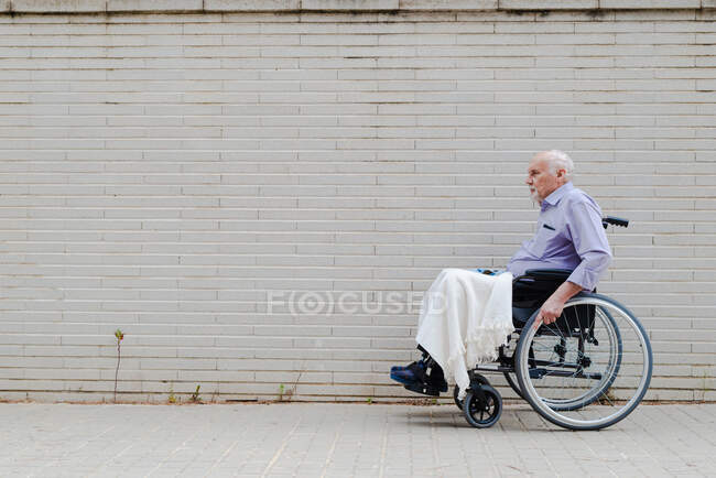 Side view of gray haired male in wheelchair riding along pavement in city — стоковое фото