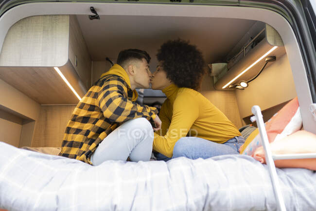 Side view of diverse man and woman kissing each other while sitting on bed in modern van during road trip — Stock Photo