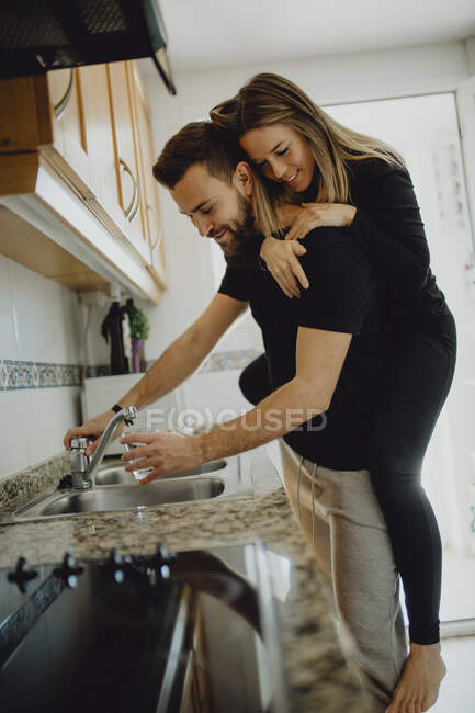 Homme barbu joyeux étreignant femme souriante tout en s'appuyant sur le placard avec évier dans la cuisine confortable à la maison — Photo de stock