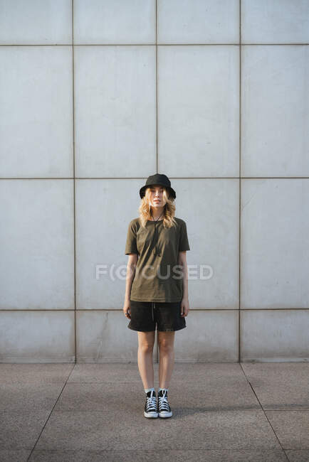 Jeune femme en vêtements décontractés regardant la caméra contre le mur de béton du bâtiment moderne sur la chaussée urbaine en journée — Photo de stock