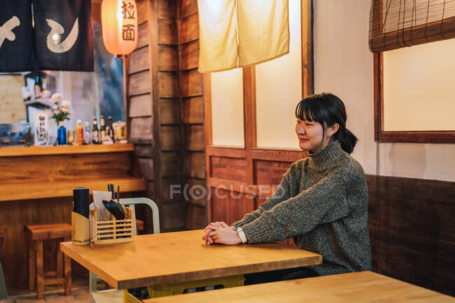 Содержание Азиатская женщина в случайном свитере глядя прочь с зубастой улыбкой, сидя за деревянным столом в рамэн-баре — стоковое фото