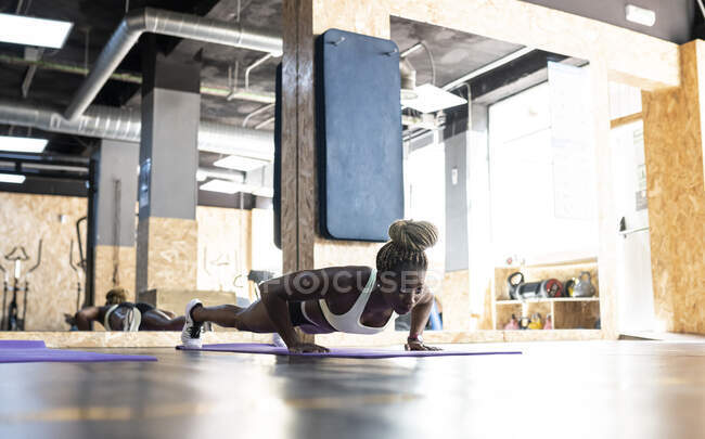 Ground level of young concentrated African American female athlete working out on mat while looking forward against mirror in gymnasium — Stock Photo